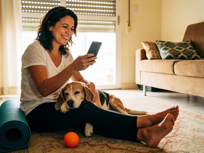 Responsável sorrindo em casa, sentada no tapete com o beagle no colo, olhando o app no celular.